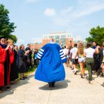 A man wearing blue academic regalia walks away from the camera with the robes billowing in the wind