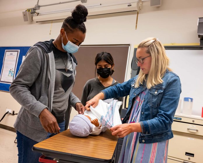 two high school students look on while a nursing instructor handles a demonstration mannequin