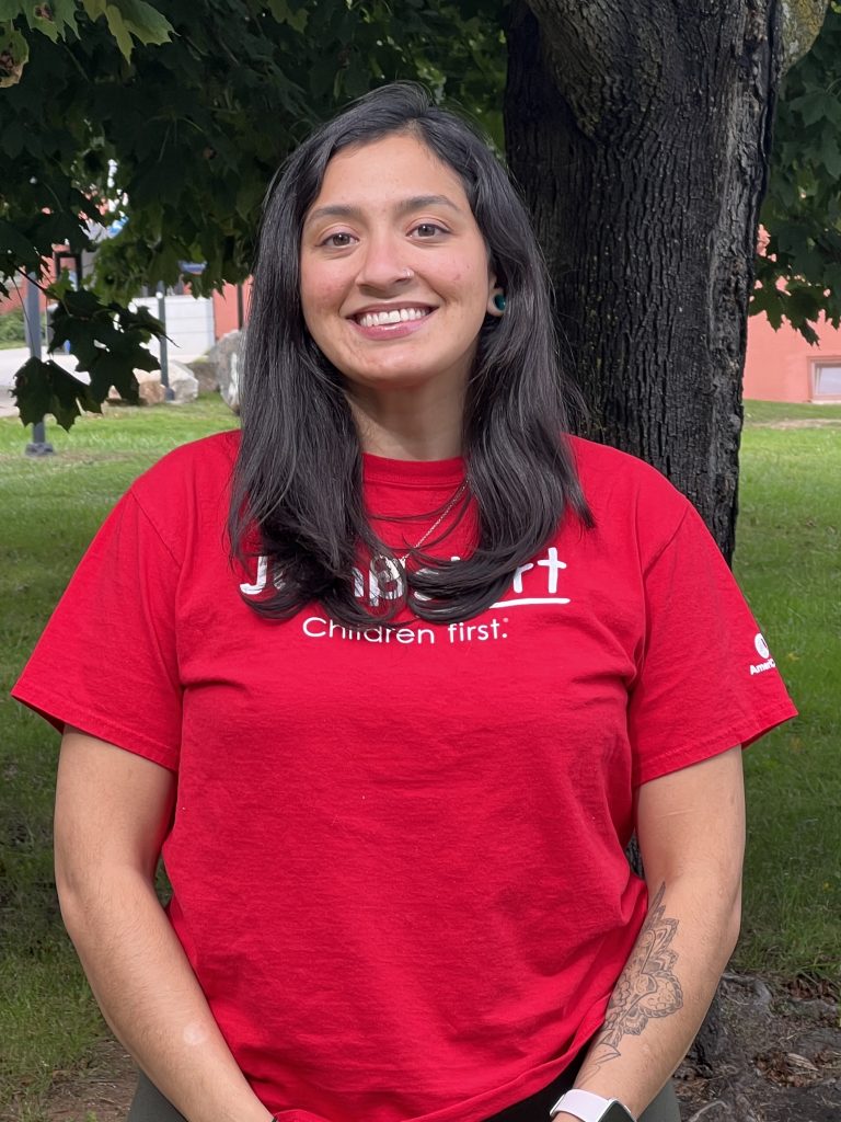 Photo of girl in red shirt standing in front of a tree smiling at the camera. 