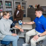 Man taking a woman's central blood pressure using an arm cuff; professor overlooks.