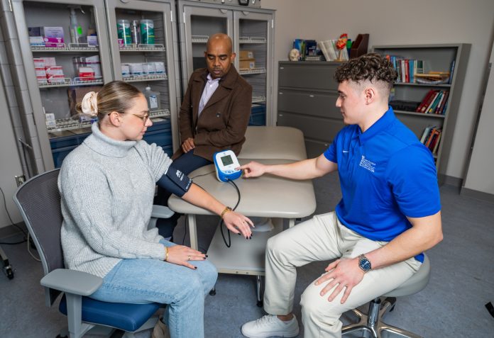 Man taking a woman's central blood pressure using an arm cuff; professor overlooks.