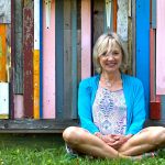 The author sits cross legged in front of a colorful wood-slab wall.