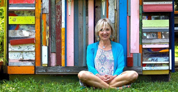 The author sits cross legged in front of a colorful wood-slab wall.