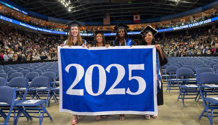 Four graduates from the Southern Connecticut State University Class of 2025 stand proudly in graduation caps and gowns, holding a large blue banner that reads “2025” inside a crowded arena filled with guests and rows of empty chairs. A digital banner above them reads “WELCOME CLASS OF 2025.”