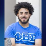 A young man stands confidently with arms crossed, smiling at the camera. He wears a bright blue Phi Beta Sigma fraternity T-shirt, and his tattooed arm reads “ONLY THE STRONG SURVIVE.” He has a full beard and natural curly hair, and the background is softly blurred with a blue gradient framing the image.