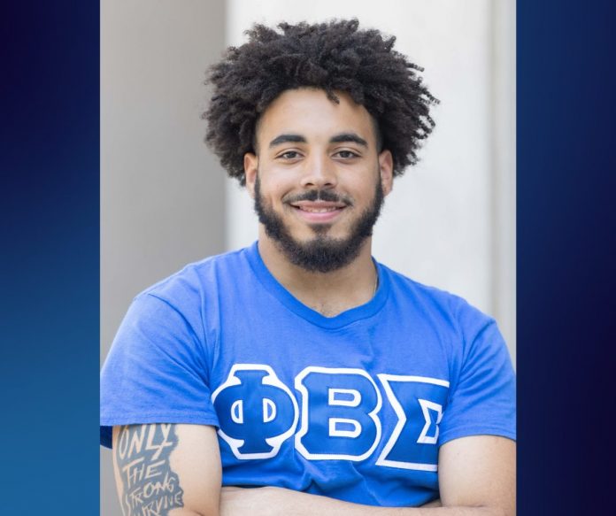 A young man stands confidently with arms crossed, smiling at the camera. He wears a bright blue Phi Beta Sigma fraternity T-shirt, and his tattooed arm reads “ONLY THE STRONG SURVIVE.” He has a full beard and natural curly hair, and the background is softly blurred with a blue gradient framing the image.