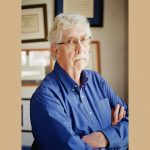 Dr. Fred Volkmar, a distinguished older man with white hair, glasses, and a mustache, stands with arms crossed wearing a blue button-down shirt. He is positioned in front of a wall decorated with framed certificates and awards in his office.