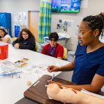 A nursing instructor in navy blue scrubs demonstrates how to insert an IV using a medical training arm, while a group of attentive high school students watch during a hands-on learning session in a clinical simulation lab.