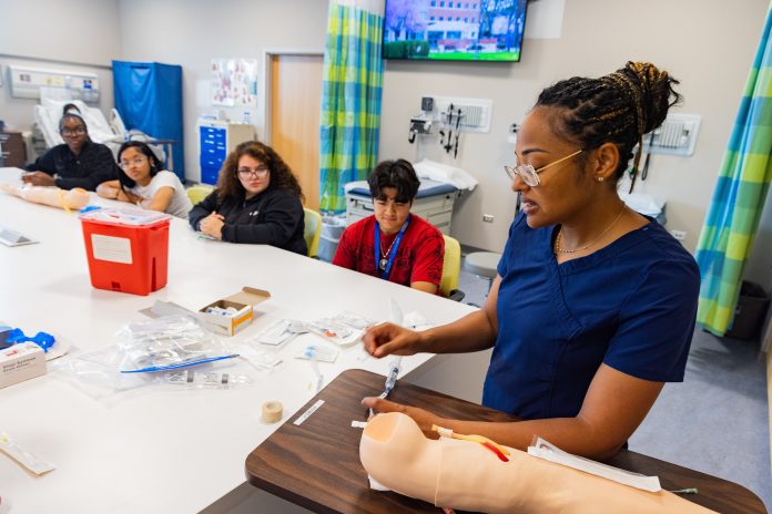 A nursing instructor in navy blue scrubs demonstrates how to insert an IV using a medical training arm, while a group of attentive high school students watch during a hands-on learning session in a clinical simulation lab.