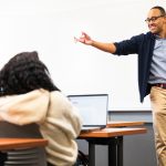 Dr. Jonathan Wharton gestures while teaching a political science class at Southern Connecticut State University, engaging students seated at desks with laptops in a brightly lit classroom.