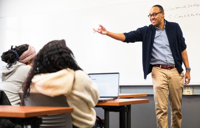 Dr. Jonathan Wharton gestures while teaching a political science class at Southern Connecticut State University, engaging students seated at desks with laptops in a brightly lit classroom.