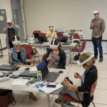 A group of adult learners seated and standing in a classroom wearing white virtual reality headsets and holding VR controllers. Laptops, notebooks, and power cords are spread across the tables, indicating an interactive training session.