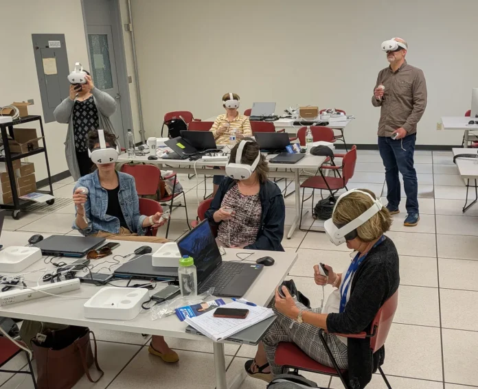 A group of adult learners seated and standing in a classroom wearing white virtual reality headsets and holding VR controllers. Laptops, notebooks, and power cords are spread across the tables, indicating an interactive training session.