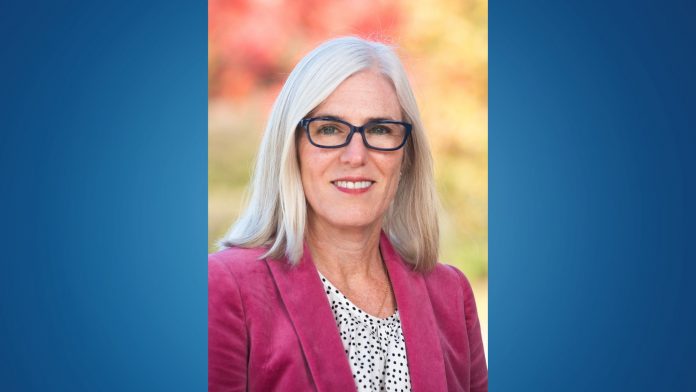 Portrait of a woman with straight gray hair wearing dark-rimmed glasses, a magenta blazer, and a white blouse with black polka dots. The background is softly blurred with autumn colors.