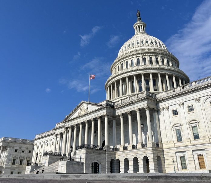 The United States Capitol building in Washington, D.C., with a clear blue sky in the background. The American flag is visible flying above the entrance, and the iconic dome with the Statue of Freedom is prominently featured.