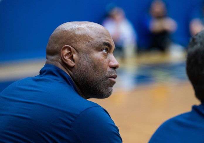 Scott Burrell, head coach of Southern Connecticut State University’s men’s basketball team, looks on intently during a game or practice. He is wearing a navy blue shirt, and the background shows a blurred gymnasium setting.