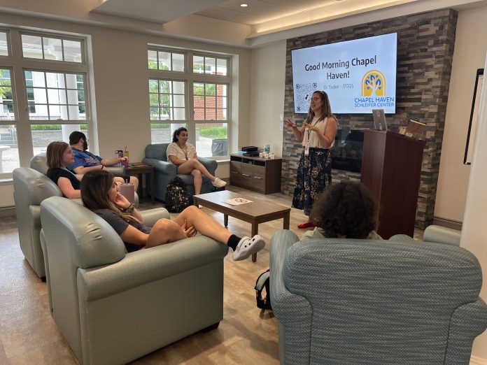 Dr. Lauren Tucker stands in front of a screen that reads “Good Morning Chapel Haven!” while speaking to a small group seated in armchairs during an assistive technology training session at Chapel Haven Schleifer Center.
