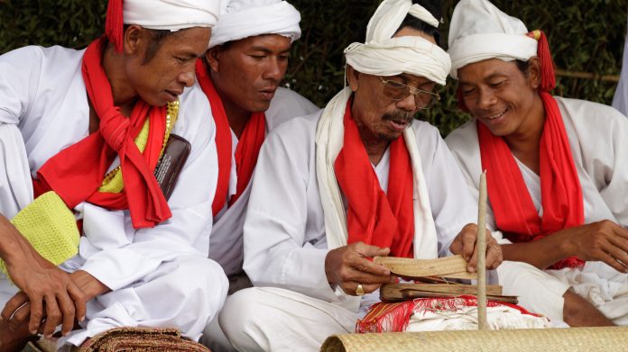 The photo depicts four white robed religious dignitaries seated on the ground. They are reading spiritual texts written on palm leaves, which one of the dignitaries is holding.