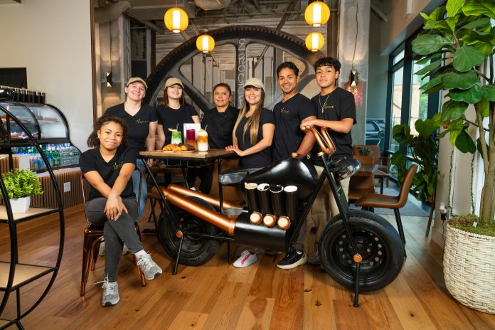 Dressed in black t-shirts, owners and staff members surround a unique display the resembles a motorcycle. A painting of a large metal wheel is in the background.