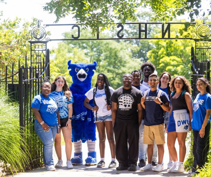 A group of Southern Connecticut State University students stand together with the school’s blue owl mascot, Otus, at the Founders Gate on campus. Everyone is smiling and wearing SCSU gear, representing school spirit and community.