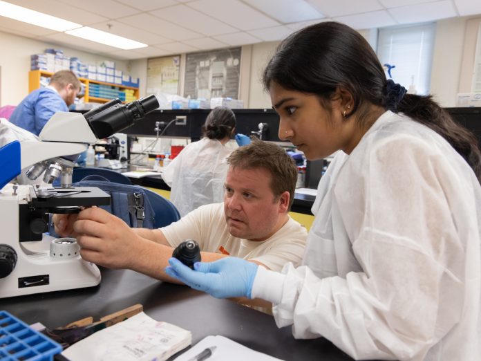 In a lab, a professor works with a student researcher at a microscope.