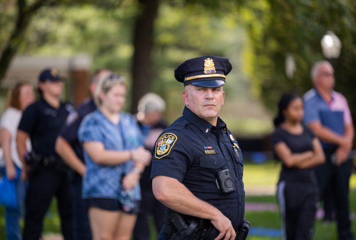 Lt. Rich Anderson, a uniformed police officer with Southern Connecticut State University, stands in sharp focus at the forefront of a campus gathering. Wearing a police cap and a body camera, he looks solemnly ahead while students and colleagues stand in the background, slightly blurred, some with heads bowed. The setting is outdoors, surrounded by trees, suggesting a moment of remembrance or ceremony.