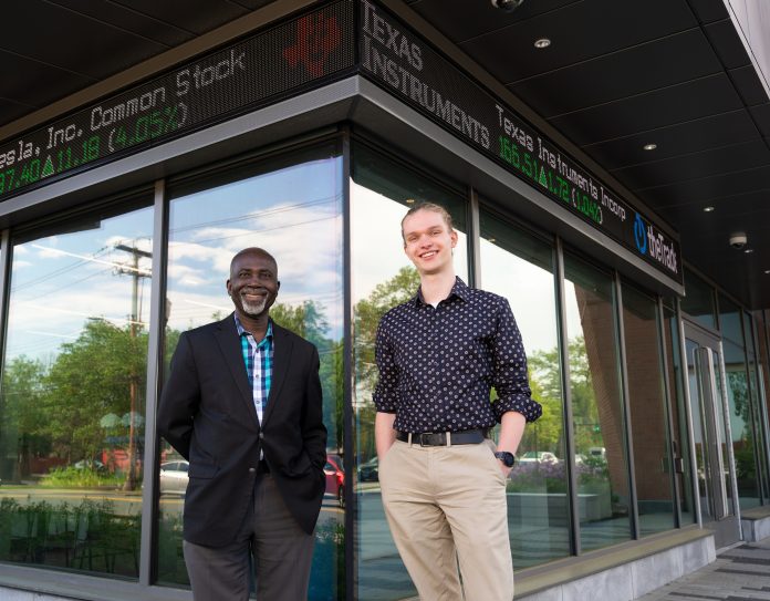 International student Tristan Janning (right) and Samual Andoh, professor of economics stand in front of the new home of the School of Business. A stock ticker tape runs above them.