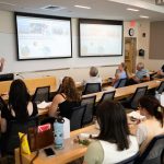 Dr. Emma Cross presents marine research findings to a room of faculty and attendees during the Werth Luncheon with Save the Sound, with slides about biodiversity and fjord water quality displayed on two large screens.