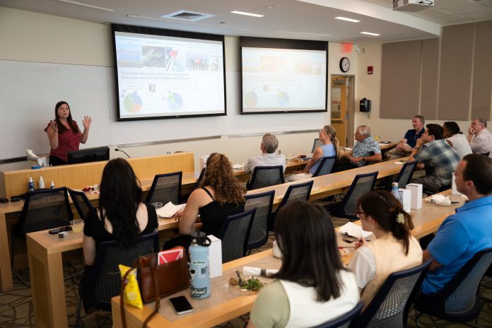 Dr. Emma Cross presents marine research findings to a room of faculty and attendees during the Werth Luncheon with Save the Sound, with slides about biodiversity and fjord water quality displayed on two large screens.