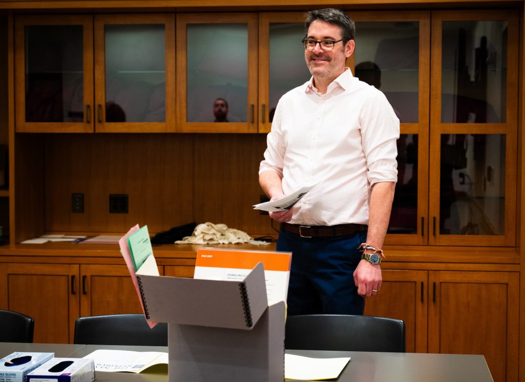 Jason Smith stands in a classroom holding papers while speaking to students near archival materials.