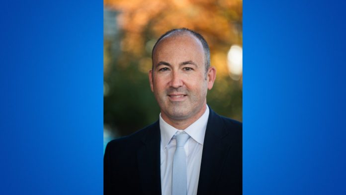 A professional headshot of Dr. Craig Hlavac wearing a dark suit, white shirt, and light-colored tie, smiling slightly while standing outdoors with a softly blurred background of autumn foliage.