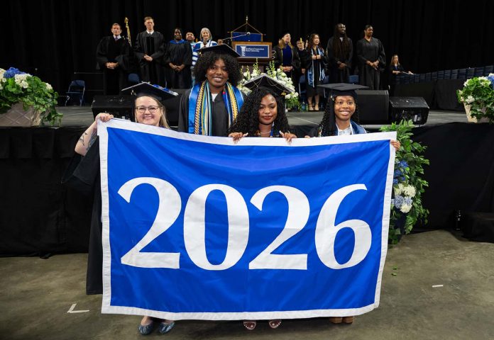 four students in graduation caps and gowns hold up a royal blue banner with large blue numerals 