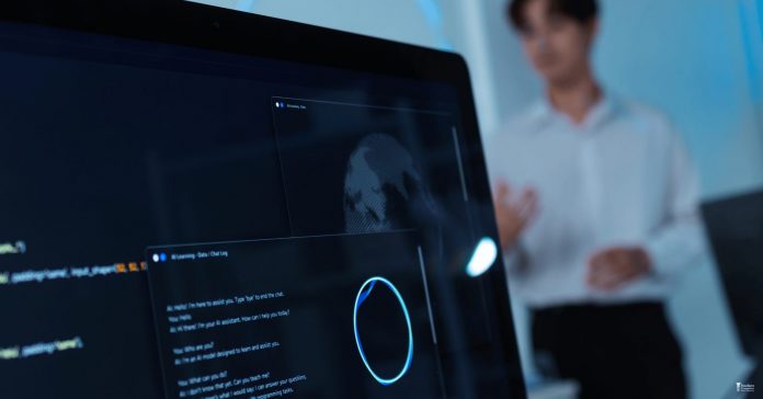 A computer screen displays an artificial intelligence chatbot interface and code, while a person stands blurred in the background. The image illustrates the growing role of AI technologies in education, workforce development, and everyday digital interactions.