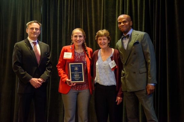 Dr. Christine Caragianis Broadbridge, second from left, stands on stage holding a Distinguished Service Award plaque alongside three colleagues during an awards ceremony. Broadbridge is wearing a red blazer and smiling as the group poses for a photo in front of a black curtain backdrop.