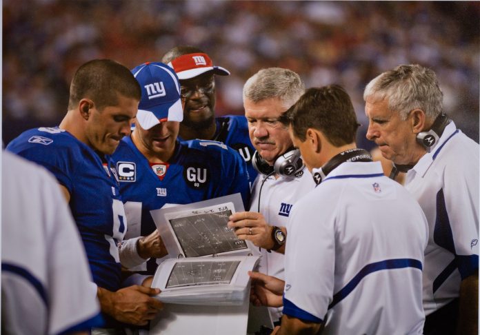In the middle of a game, New York Giants coaches and football players review a notebook of plays
