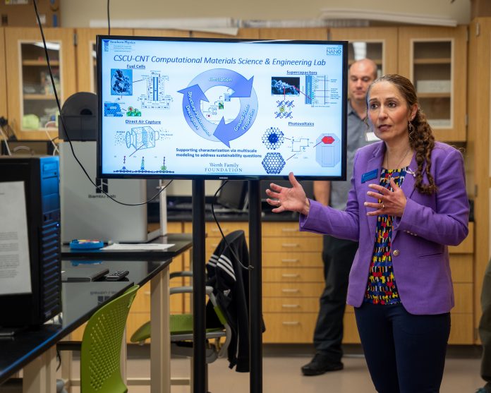 Alt Text: Dr. Christine Caragianis Broadbridge speaks in a laboratory at Southern Connecticut State University while presenting research on computational materials science and engineering. Standing beside a large display screen showing scientific diagrams and sustainability applications, she gestures as she explains the work to attendees.