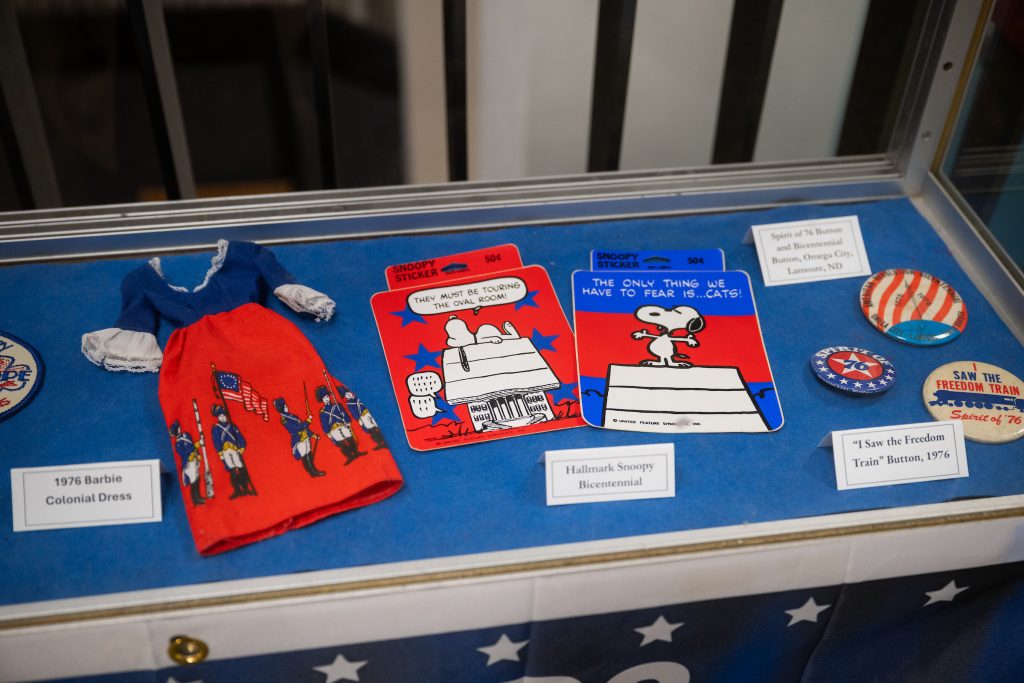 1976 items on display include red, white, and blue doll's dress, Snoopy stickers, and buttons.