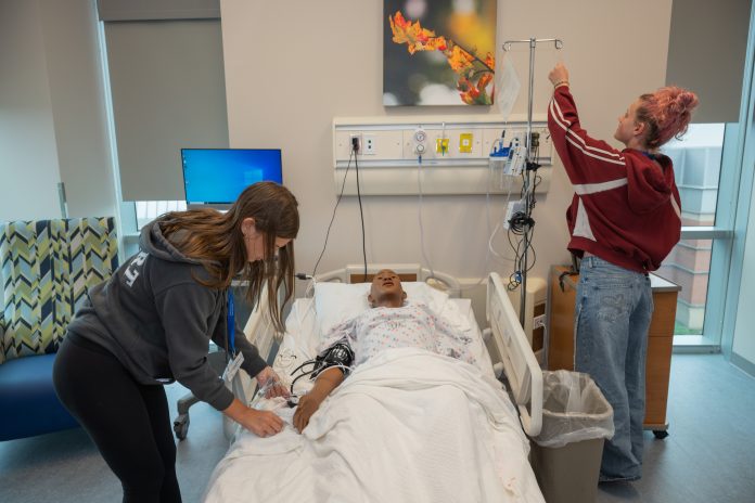 Two high school students practice patient care in a nursing simulation lab at Southern Connecticut State University, with one student adjusting an IV pole while another works beside a lifelike patient simulator in a hospital bed.