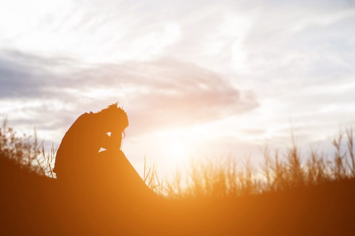 Silhouette of a person sitting alone with their head bowed while overlooking a sunset, surrounded by tall grass beneath a cloudy sky. The warm golden light creates a reflective, somber atmosphere.