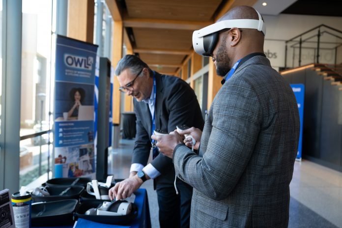 An attendee wearing a virtual reality headset holds controllers while another person assists at a technology demonstration table during Southern’s 2025 Emerging Technologies Forum.
