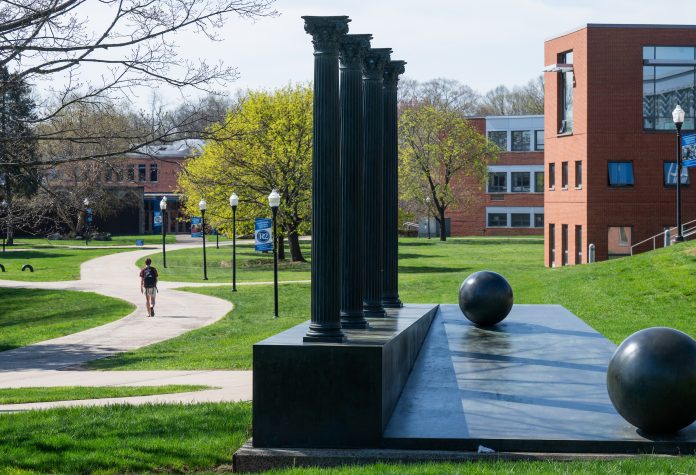 A student walks along a pathway on Southern Connecticut State University’s campus near the four-column sculpture, with academic buildings and trees in the background.