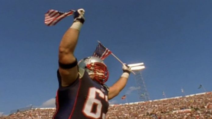 Alt text: Joe Andruzzi runs onto the football field in a New England Patriots uniform, raising two American flags above his head.