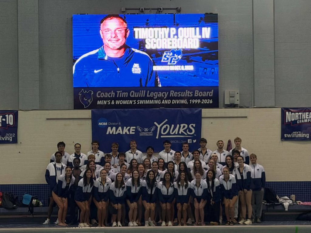 Southern Connecticut State University men’s and women’s swimming and diving teams pose beneath the Timothy P. Quill IV Scoreboard at Hutchinson Natatorium following its dedication in his memory.
