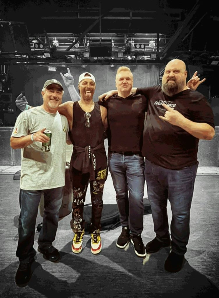 Four longtime friends stand arm in arm in front of a stage during an April 2025 benefit concert for Darik Pearson and his family at Toad’s Place in New Haven.